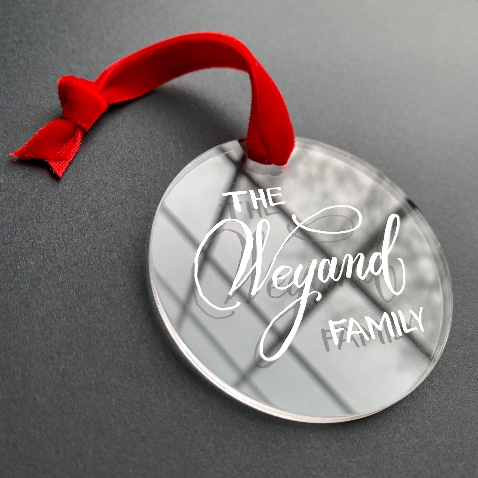 Clear round acrylic ornament hand-lettered 'The Weyand Family' with a red ribbon