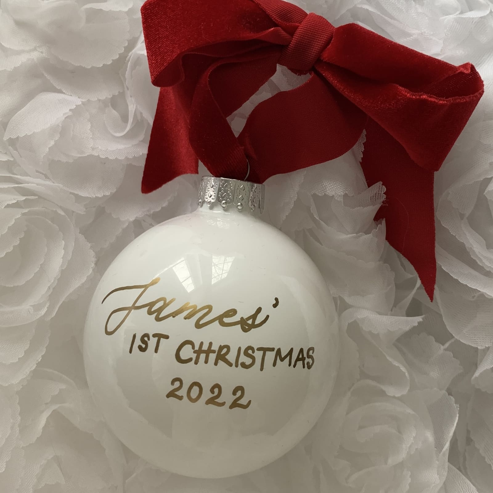 White glass ball ornament hand-lettered 'James' 1st Christmas 2022' in gold with a red velvet bow