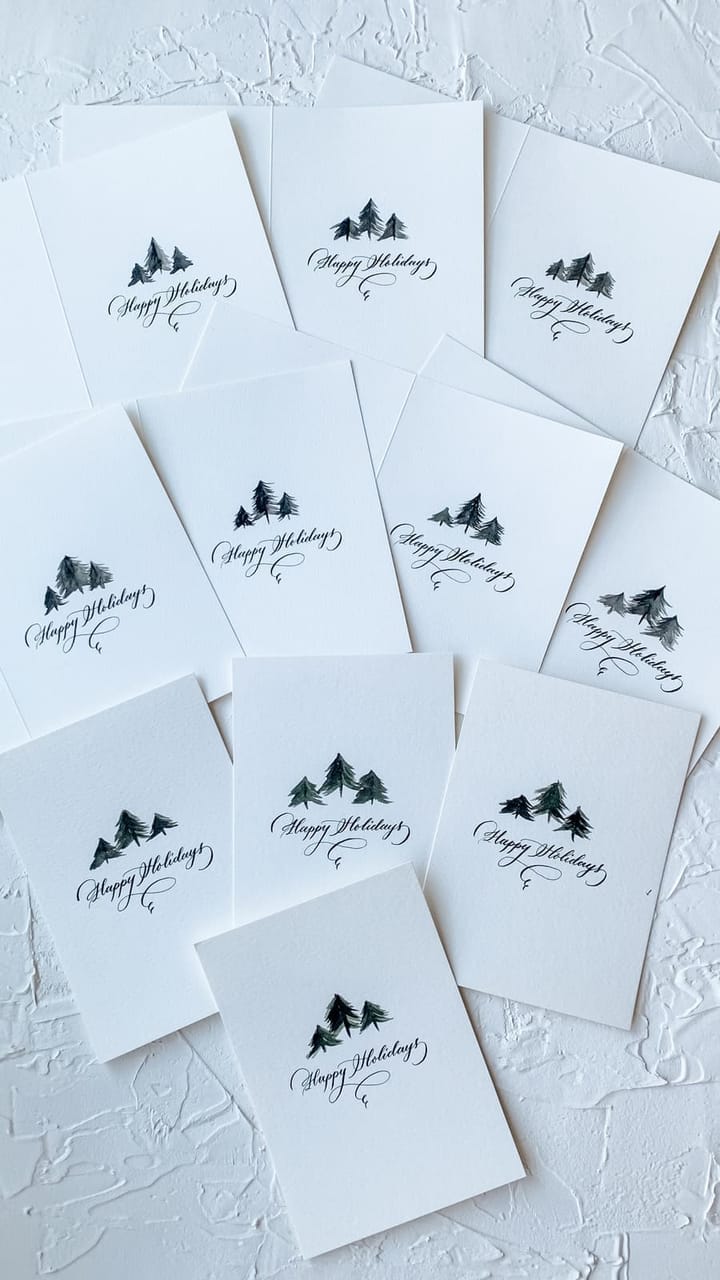 A stack of Happy Holidays cards hand-lettered with painted pine tree illustrations