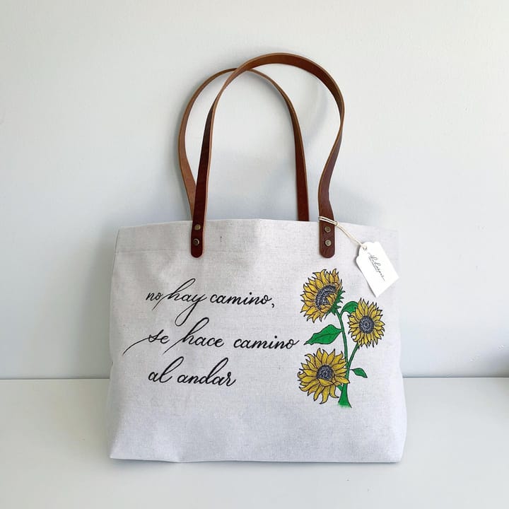Canvas tote bag hand-lettered 'no hay camino, se hace camino al andar' beside a painted sunflower illustration