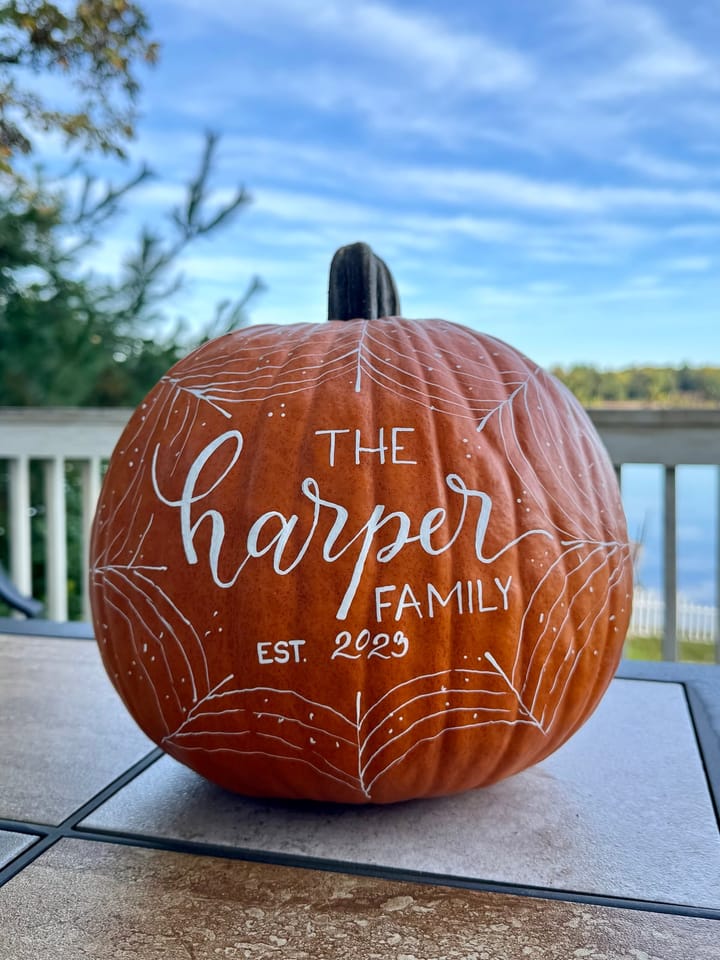 A hand-lettered pumpkin reading The Harper Family, Est. 2023, with a white spiderweb border, on a deck with a lake view
